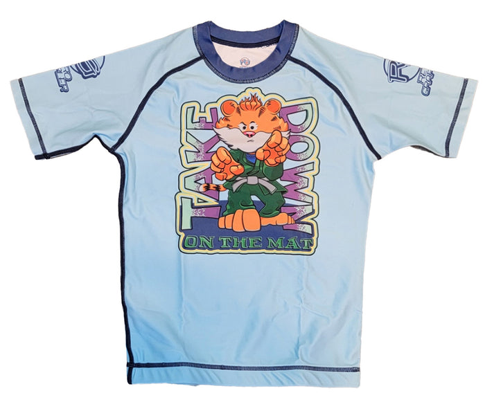 The Retro Grappler Youth - Take Down Tiger rash guard is light blue with short sleeves, featuring a cartoon tiger in green and TAKE DOWN ON THE MAT text. Made from four-way stretch fabric for comfort and mobility.